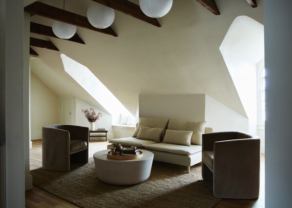 A modern loft bedroom with a sloped ceiling, a large window, and a cozy seating area with a sofa and two armchairs.
