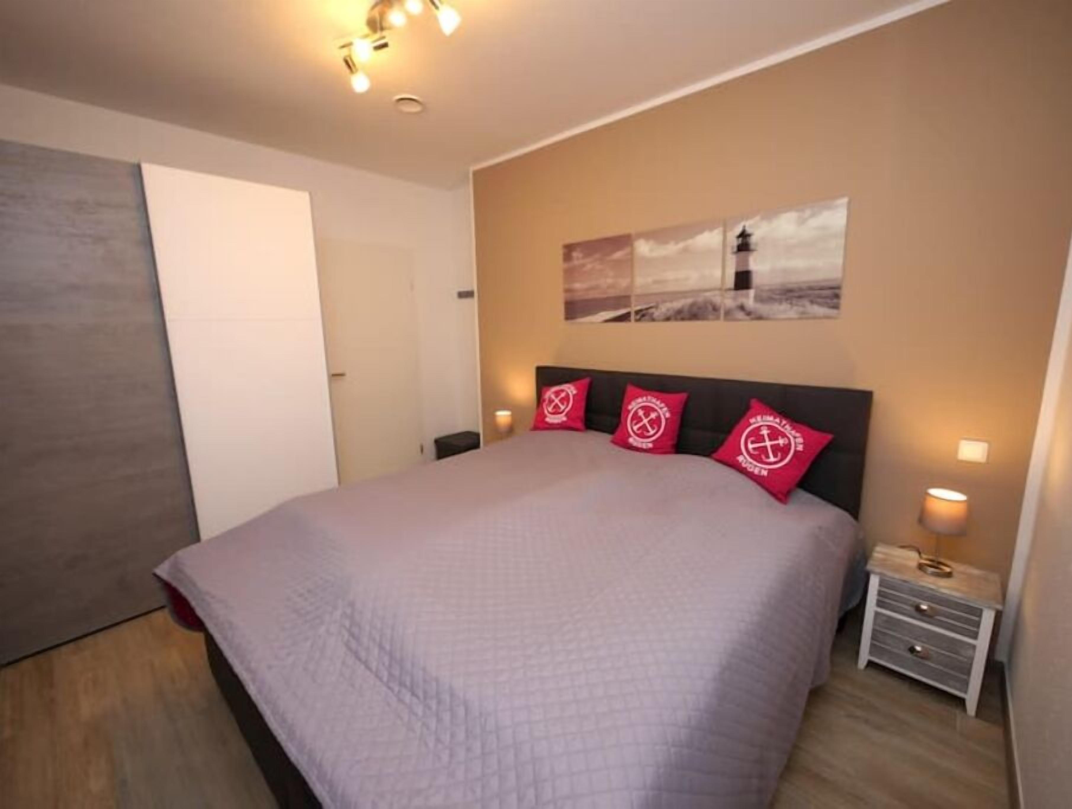 A bedroom with a large bed, a nightstand, a wall-mounted lamp, and a painting of a lighthouse.