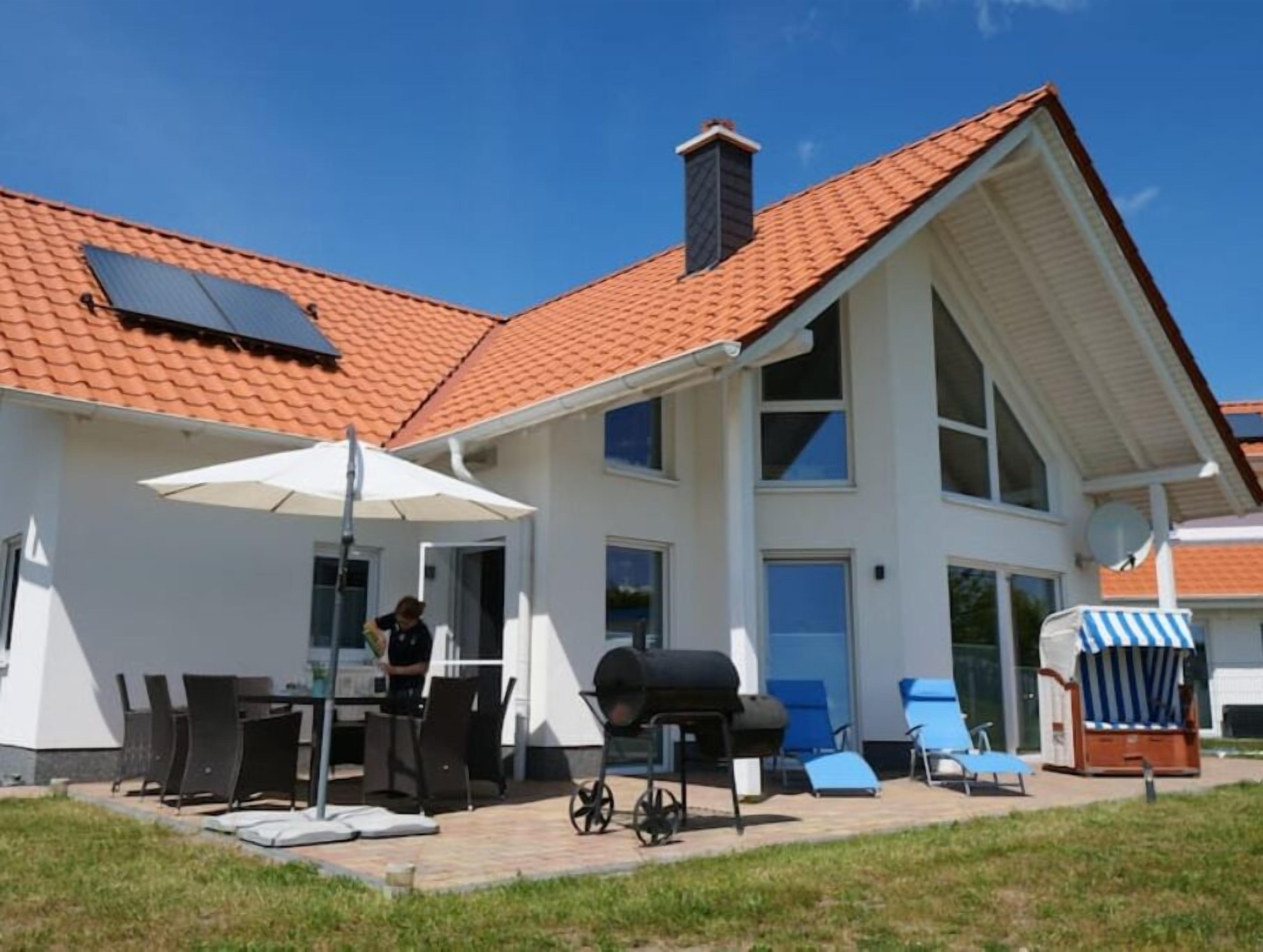 Una casa con techo de tejas rojas, un patio con mesa y sillas, una parrilla y una silla de playa a rayas.