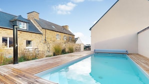 A swimming pool with a wooden deck, stone buildings, and a clear sky.