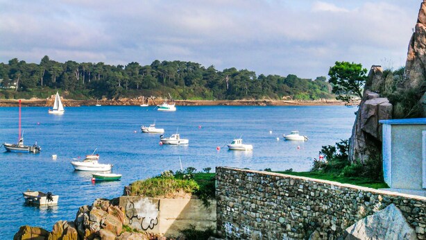 A coastal scene with boats on calm waters, a stone wall, and a rocky cliff.