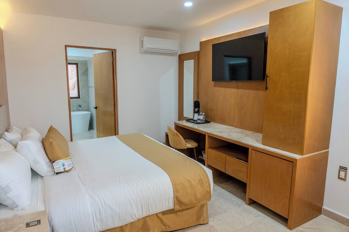 A hotel room with a bed, a desk with a chair, a television mounted on the wall, and an air conditioning unit.