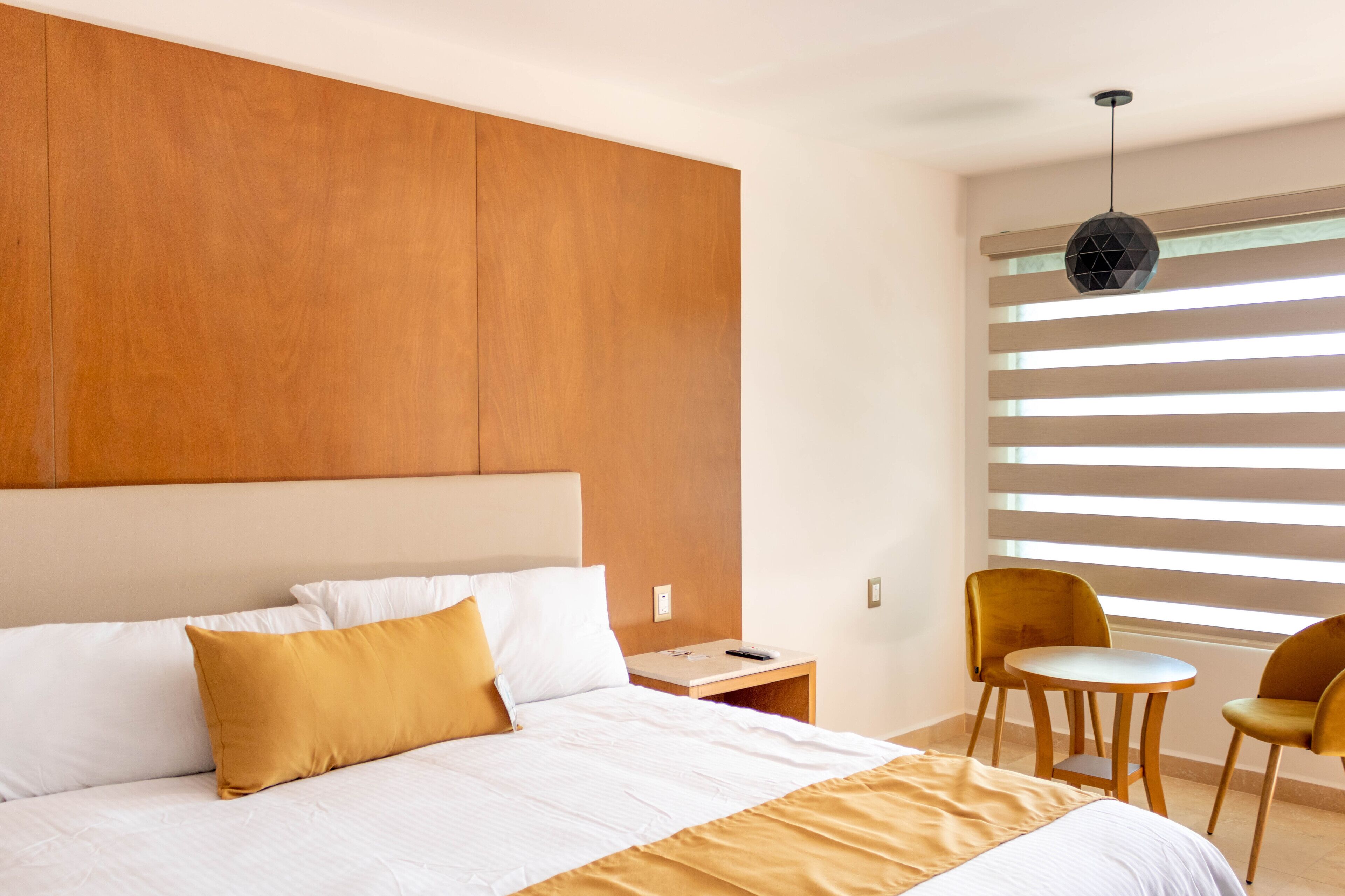 A modern hotel room with a large bed, a nightstand, a small table, two chairs, and a window with blinds.