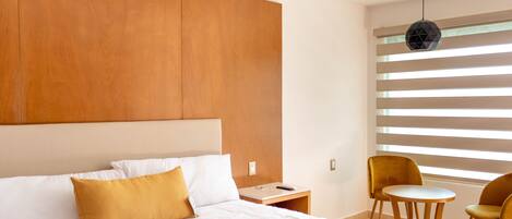 A modern hotel room with a large bed, a nightstand, a small table, two chairs, and a window with blinds.