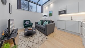 A modern living room with a grey sofa, a black coffee table, and a green armchair. A large window provides natural light, and a kitchen is visible in the background. - SERCASA - Appletree Court - Kingston (Kingston Upon Thames)