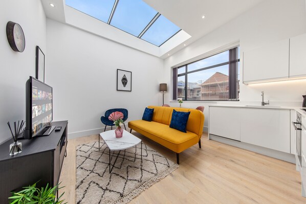 A modern living room with a sofa, a coffee table, and a TV. There is a skylight above the sofa and a window with a view of buildings. - SERCASA - Appletree Court - Kingston (Kingston Upon Thames)