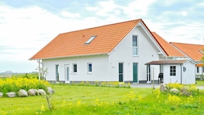 A white house with a red-tiled roof, a small porch, and a well-maintained garden with yellow flowers and rocks.