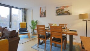 A modern living room with a dining area, featuring a large window, a sofa, a dining table with chairs, and a potted plant. - Fabulous 3 bedroom property in city centre location. Sleeps 5. (Edinburgh)