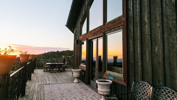 A wooden deck with a table and chairs, large potted plants, and a view of the sunset.