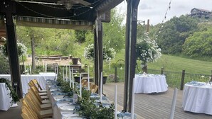 An outdoor dining area with long wooden tables set with white tablecloths, blue napkins, and silverware.