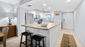 A modern kitchen with white cabinets, a central island with stools, and a dining area with a table and chairs. - Simply step across the street from this condo to reach Florida's premier beach! (Siesta Key)
