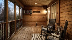 Eine hölzerne Veranda mit Deckenventilator, Dartscheibe und einem Dartspiel an der Wand. Zwei Stühle mit Kissen und ein gemustertes Teppich sorgen für Komfort.