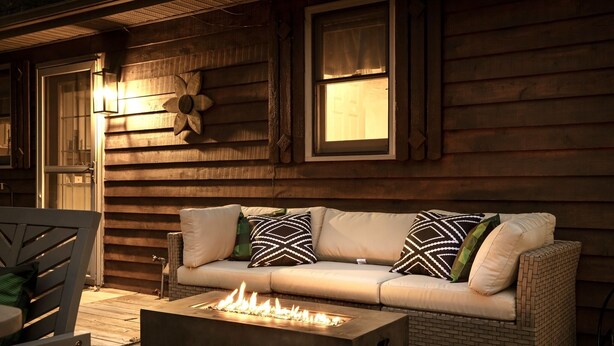 Ein gemütlicher Außenbereich mit Feuerschale, einer gepolsterten Couch mit gemusterten Kissen und einer Holzwand mit Fenstern.