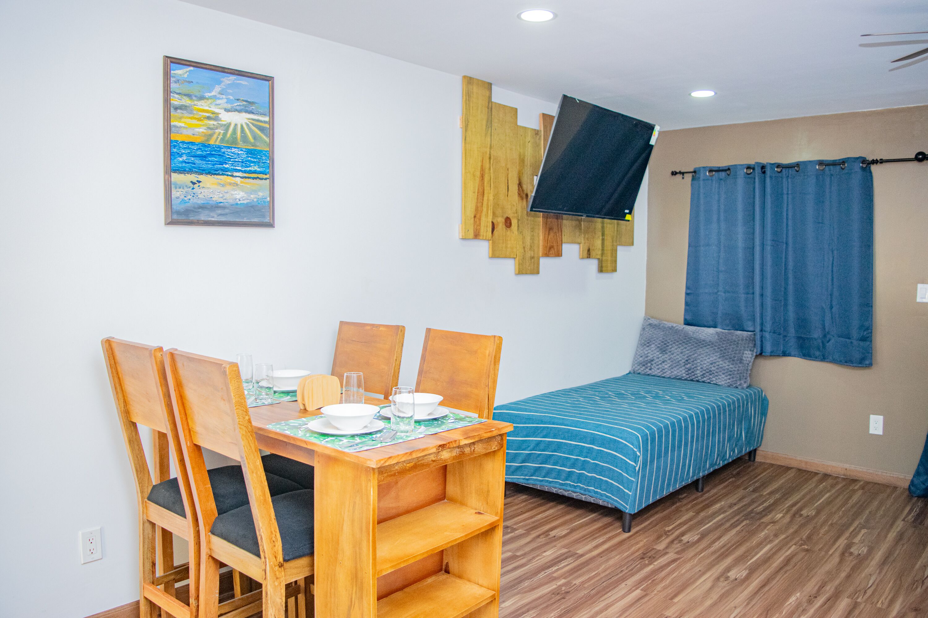 A small, well-lit room with a wooden dining table and chairs, a bed with a blue-striped bedspread, a mounted TV, and a painting of a landscape on the wall.