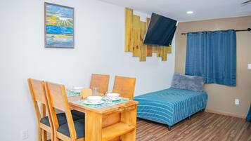 A small, well-lit room with a wooden dining table and chairs, a bed with a blue-striped bedspread, a mounted TV, and a painting of a landscape on the wall.