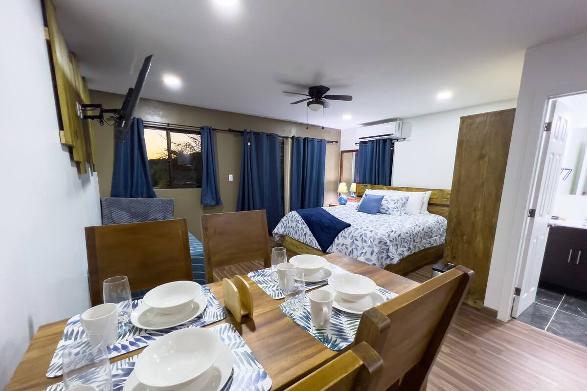 A hotel room with a dining table set for four, a bed with a patterned bedspread, a ceiling fan, and an air conditioning unit.