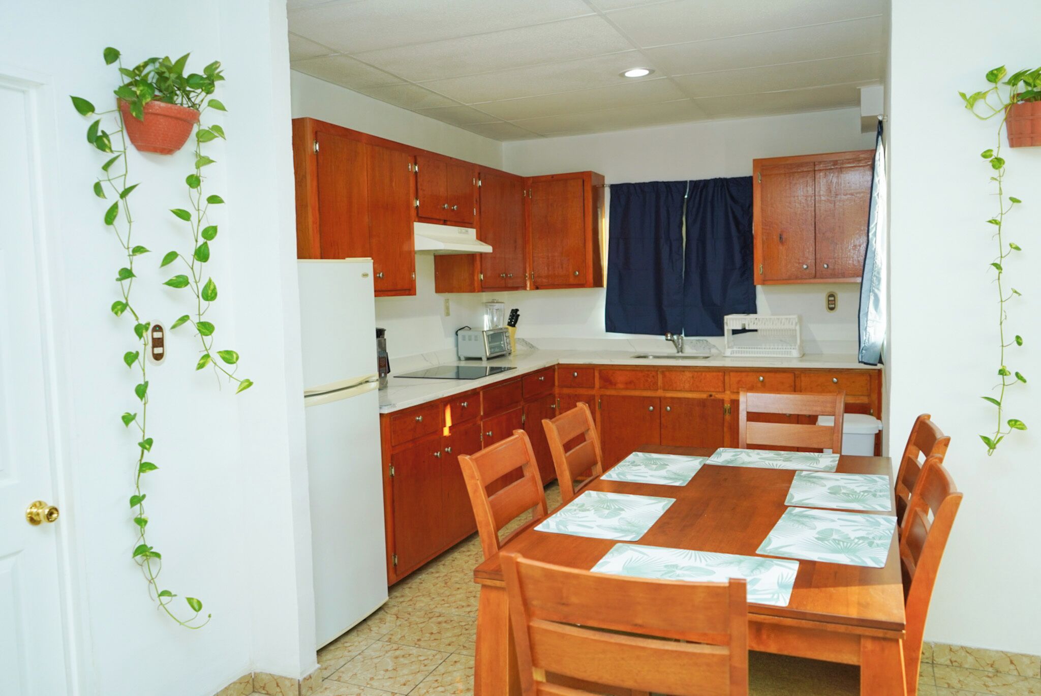A compact kitchen with wooden cabinets, a dining table with chairs, and a window with curtains.