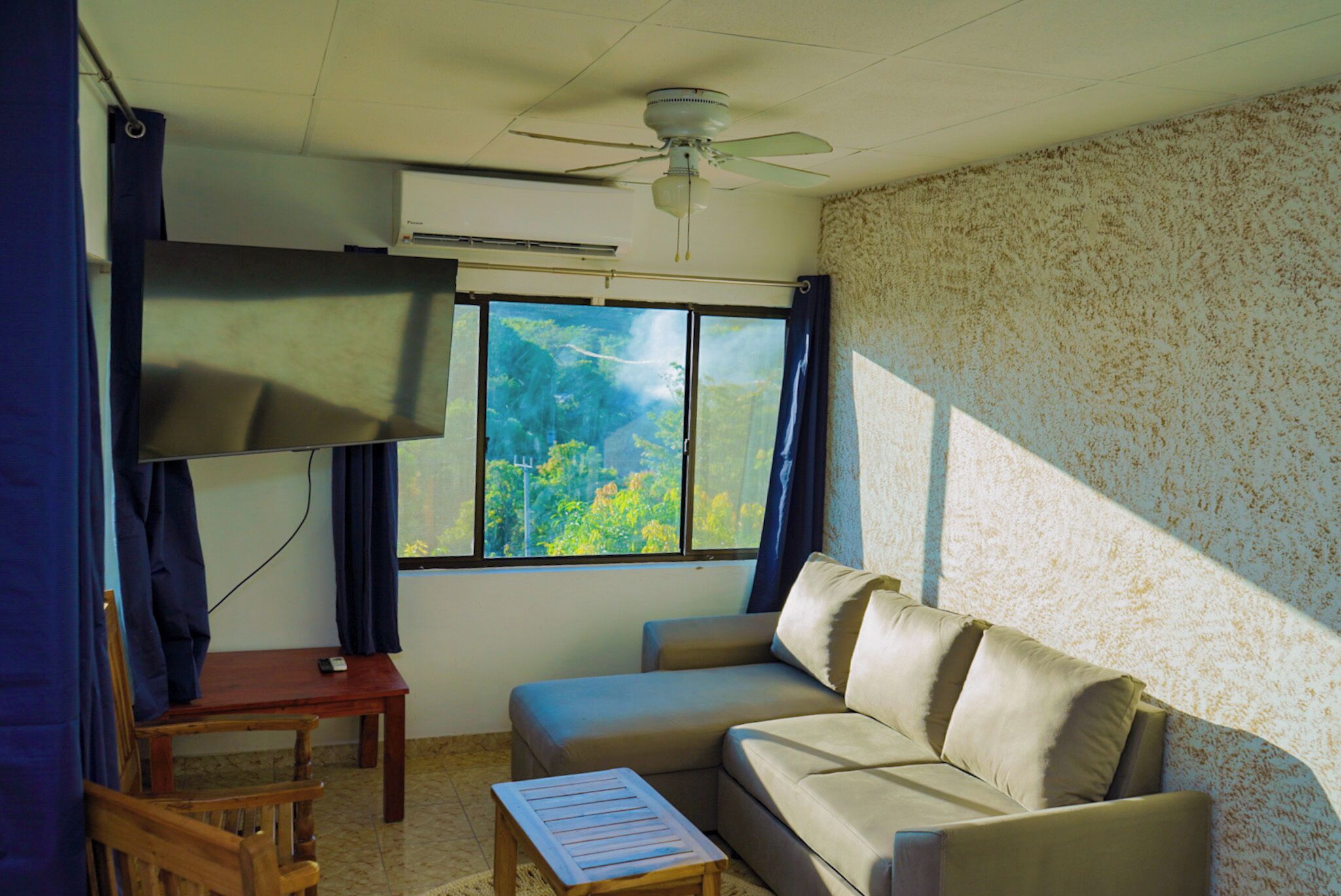 A room with a sofa, a small table, a chair, a ceiling fan, and a window with a view of greenery.
