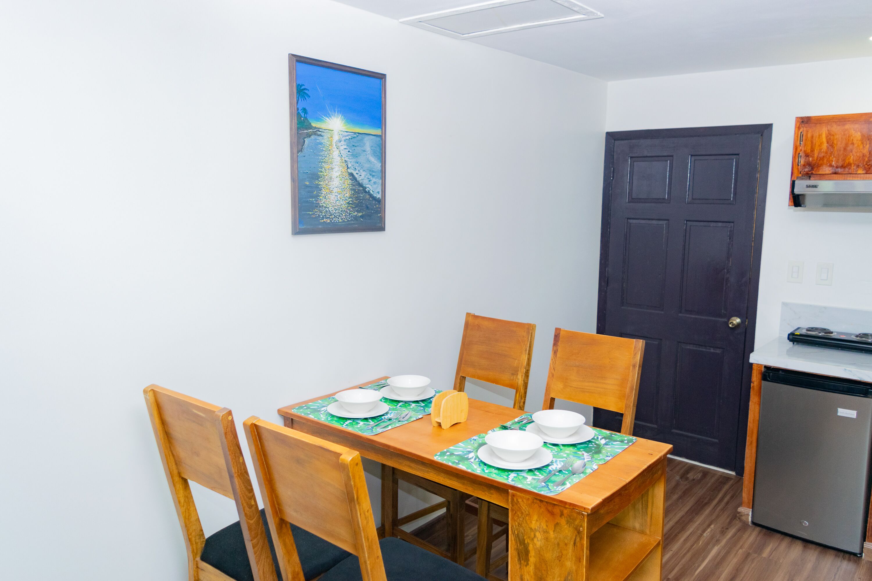 Ein Essbereich mit einem Holztisch, gedeckt für vier Personen, Stühle, einem Gemälde mit einer Strandansicht, einer dunklen Tür und einer kleinen Küche mit einem Kühlschrank.