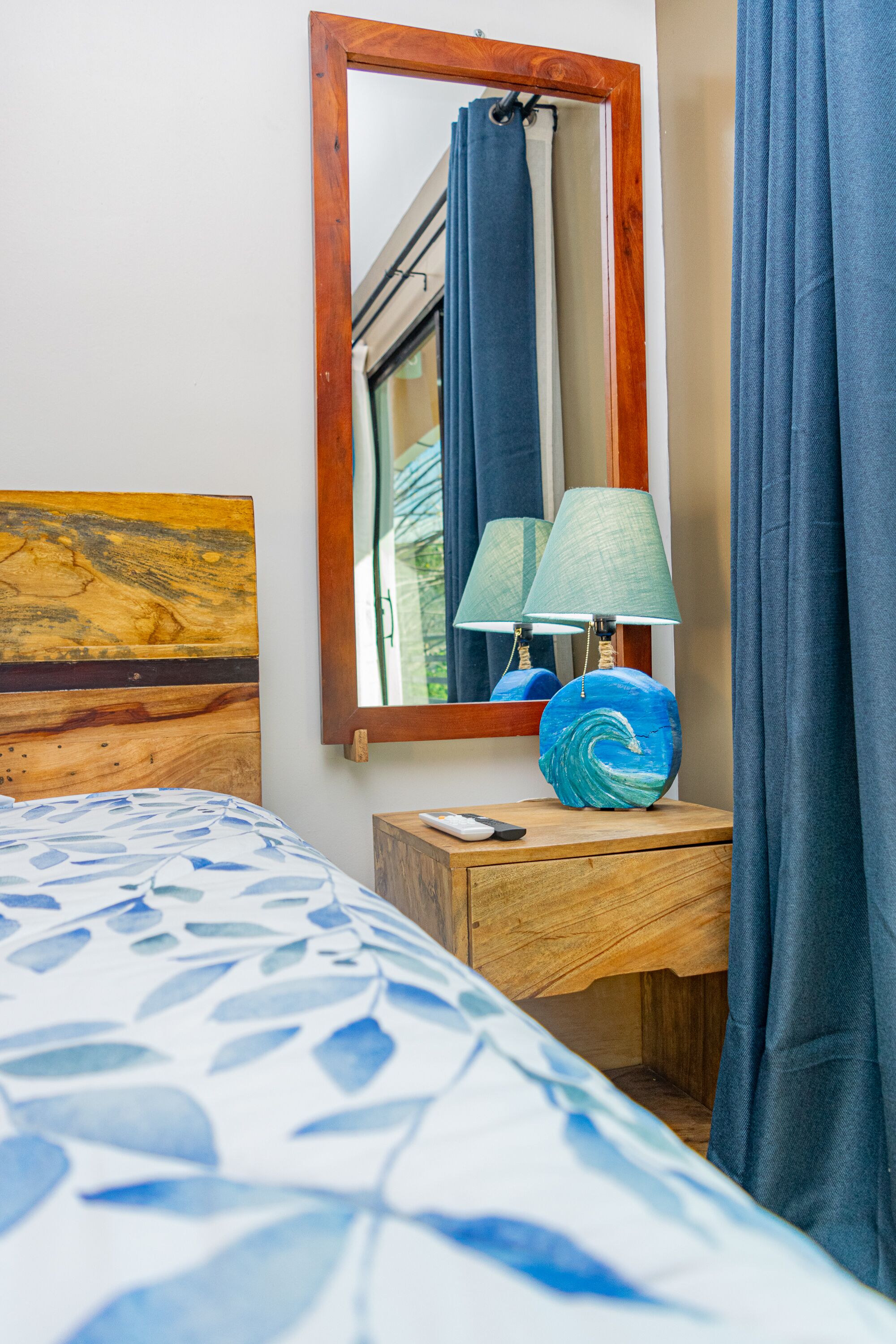 A bedroom with a bed, a wooden nightstand, a lamp, a mirror, and curtains.