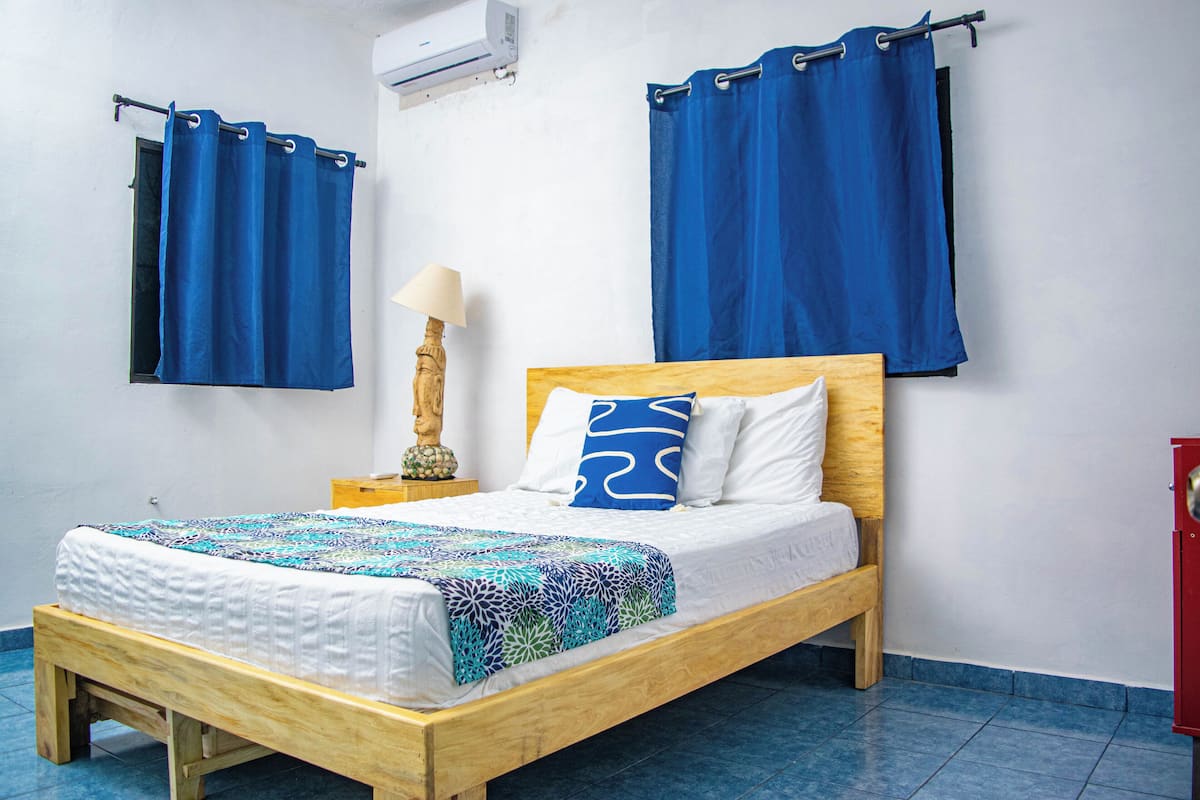 A single bed with a wooden frame, a white mattress, and a patterned blanket. There is a wooden nightstand with a lamp and a decorative item beside the bed. Blue curtains hang on the windows, and a red cabinet is visible to the right.