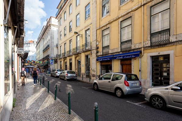A narrow street lined with yellow buildings, parked cars, and pedestrians.