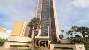 A tall, modern building with a glass facade, surrounded by palm trees and greenery. - 4 BR Oceanfront Oasis: Luxury Amidst the Bustle w Heated Pool & Private Balcony (South Padre Island)