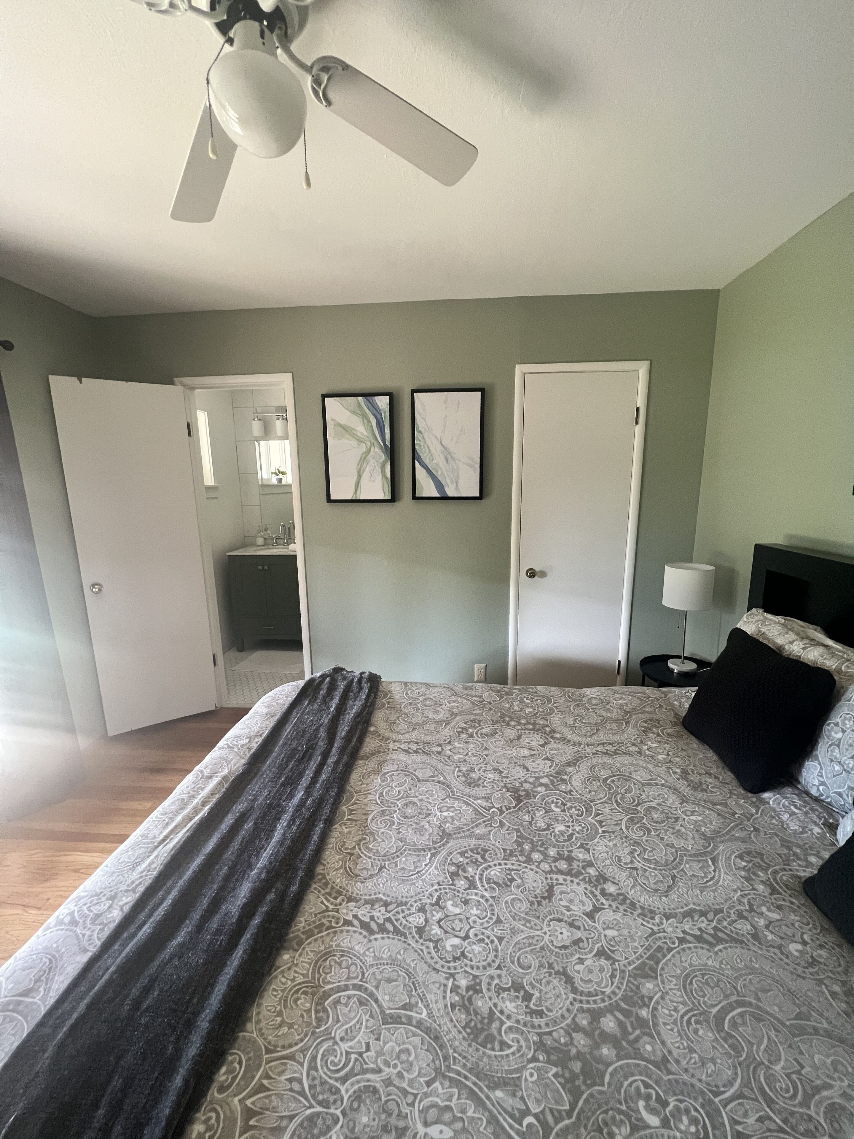 Et soveværelse med seng, ventilator, natbord og dør.