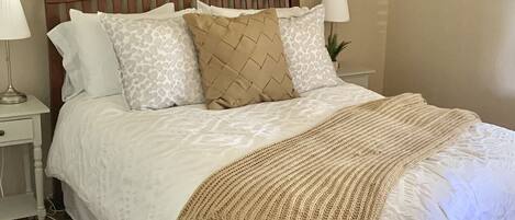 Un dormitorio con cabecera de madera, ropa de cama blanca, una manta color beige y una lámpara de pared.