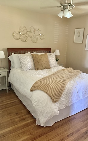 Quarto com cabeceira de madeira, roupa de cama branca, manta bege e luminária fixada na parede.