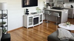 Una cocina moderna con una isla central, un comedor con dos taburetes tipo barra, un televisor de pantalla plana montado en la pared y un pequeño rincón de estar con sofá y reposapiés.