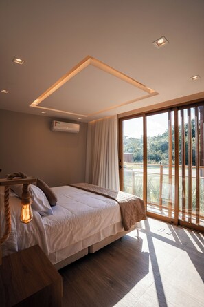 A bedroom with a bed, a wooden bedside table, a hanging light fixture, and a view of greenery through a sliding glass door. - Casa Oceano- Praia do Forte (Açu da Torre)
