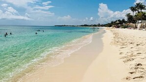Una spiaggia con acqua turchese trasparente, persone che nuotano e una riva sabbiosa con palme e ombrelloni.