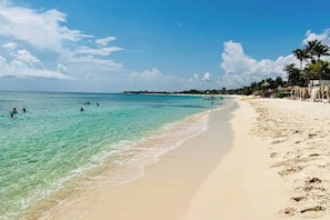 Une plage à l’eau turquoise et limpide, où l’on peut se baigner, avec une rive sablonneuse bordée de palmiers et parsemée de parasols de plage.
