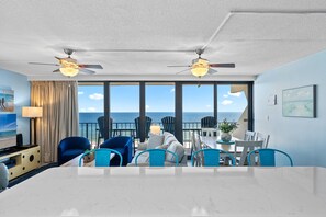 A modern hotel room with a large dining table, blue chairs, a sofa, and a balcony with ocean views.