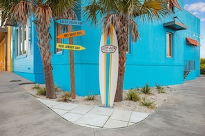 Un edificio azul con palmeras, un letrero con forma de tabla de surf y señalización que indica Cocoa Beach, Key West y Nueva Jersey.