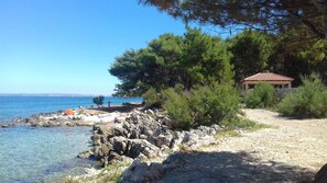 A coastal area with a rocky beach, clear blue sea, and a small building with a red-tiled roof.