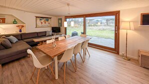 A spacious living area with a wooden dining table, white chairs, a dark sofa, and a large window offering a view of the outdoors.