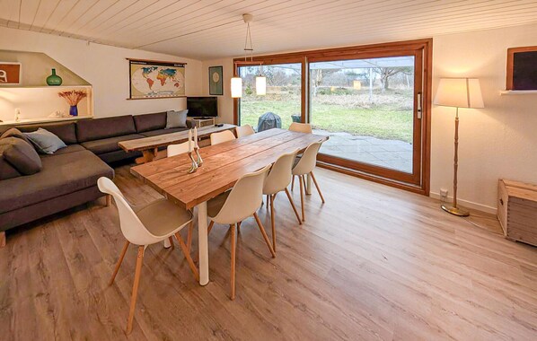 Een ruime zithoek met een houten eettafel, witte stoelen, een donkere bank en een groot raam met uitzicht op de omgeving.