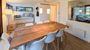 A dining area with a wooden table and white chairs, a sofa with cushions, and a window with a view.