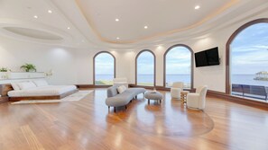 Une chambre spacieuse avec un grand lit, un coin salon avec un canapé et des fauteuils, et une vue sur la mer grâce à de grandes fenêtres.