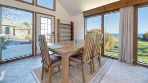 A dining area with a wooden table and chairs, large windows with curtains, and a view of the outdoors. - Bay Cottage by Avantstay Waterfront, Pier, Pond (Dameron)