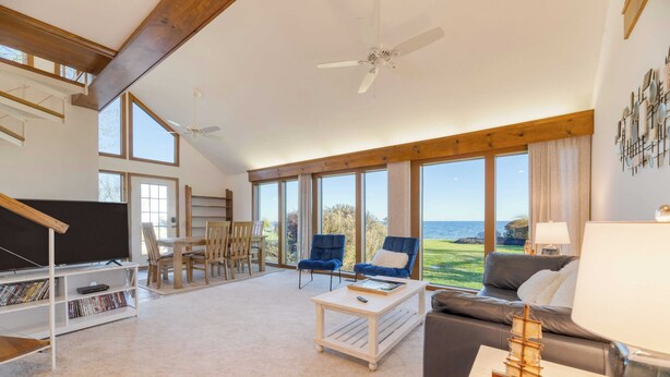 A spacious living room with a large window overlooking a scenic view, a ceiling fan, and a bookshelf filled with books. - Bay Cottage by Avantstay Waterfront, Pier, Pond (Dameron)