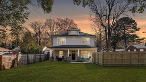 A two-story house with a front porch, a fenced backyard, and trees in the background.