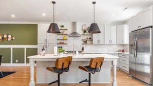 A modern kitchen with white cabinets, a central island with two bar stools, pendant lighting, and a stainless steel refrigerator.