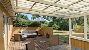 Un patio techado con piso de madera, un sofá, una mesa con una jarrón y una sombrilla.