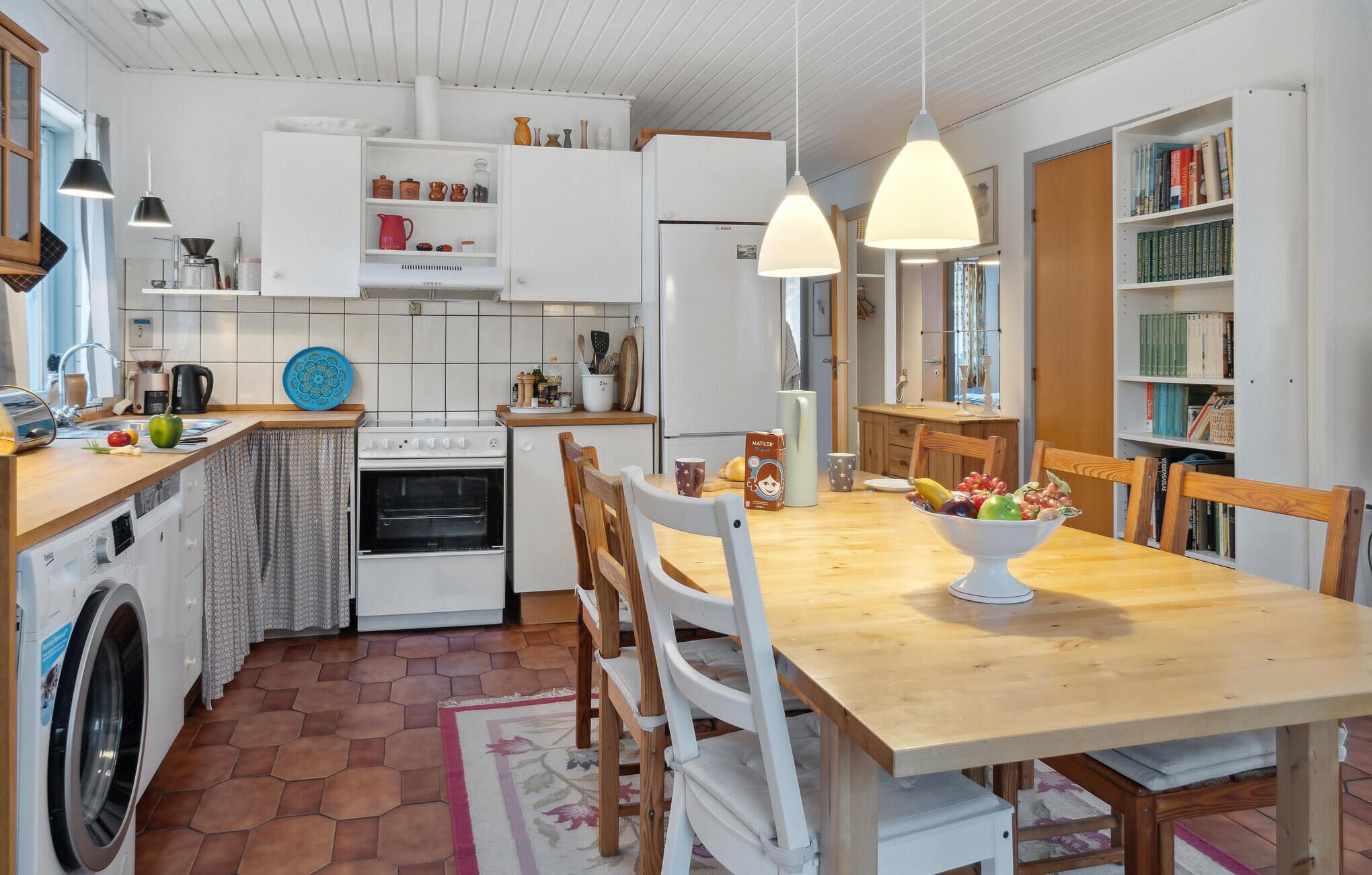 A modern kitchen with a dining area, featuring a stove, oven, washing machine, and a bookshelf filled with books.