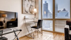 A modern hotel room with a large window offering a city view, a flat-screen TV, a desk with a chair, and a lamp. - Walk to Central Park! Cozy 1BR w. AC, Wifi & Heating (New York)