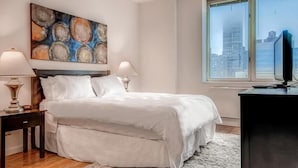 A bedroom with a large bed, two bedside tables with lamps, a painting above the bed, and a window with a view of buildings. - Walk to Central Park! Cozy 1BR w. AC, Wifi & Heating (New York)