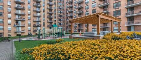 A high-rise residential building with multiple balconies, a central courtyard with a pergola, and a playground.